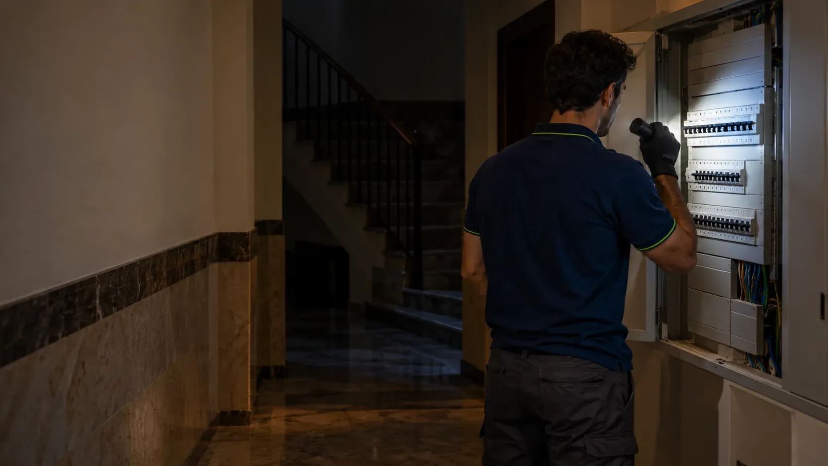 Electrician checking the communal services board of an apartment building with the staircase in darkness